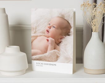 May include: A newborn baby boy is lying on a white blanket. The baby has light skin and blue eyes. The baby is wearing a white onesie. The image is a birth announcement card with the text "Lucas Davidson 28.02.2023".