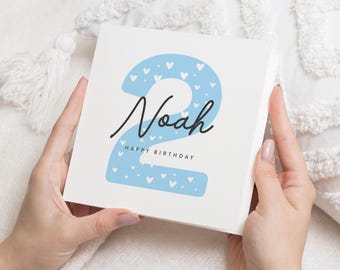May include: A white card with a blue number two decorated with white hearts. The card says "Noah" and "Happy Birthday".