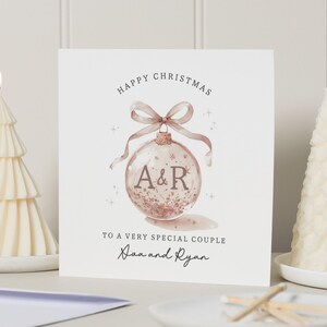 Può includere: Un biglietto di Natale con un design di ornamento ad acquerello, con le iniziali "A&R" all'interno. Il biglietto recita "Buon Natale" e "A una coppia molto speciale" con i nomi sottostanti. Due candele bianche a forma di albero accese affiancano il biglietto.