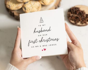May include: White card with black text that reads "To my husband on our first Christmas as Mr & Mrs Jones". A small black and white drawing of a Christmas tree is above the text. Two small red hearts are below the text.