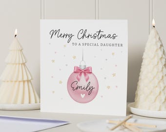 May include: A Christmas card with the message "Merry Christmas to a Special Daughter." The card features a pink ornament with a bow and the name "Emily." Two lit candles shaped like Christmas trees flank the card.