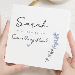 Peut inclure: Carte blanche avec le nom "Sarah" en écriture élégante. Le texte "Will you be my Something blue?" est en dessous, avec un petit cœur bleu et une illustration florale. Carte de proposition.