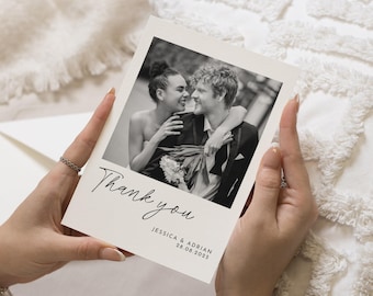 May include: A cream-colored thank you card held by a person. The card features a black and white photograph of a couple embracing. The words "Thank you" are written in elegant script, with the names "JESSICA & ADRIAN" and the date "28.06.2025" below.