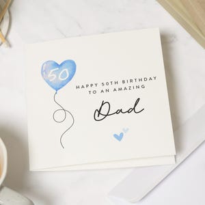 May include: A square white greeting card featuring a watercolor blue heart-shaped balloon with the number 50 inside. The text reads Happy 50th Birthday to an amazing Dad in black font, accompanied by two small blue hearts below the word Dad.