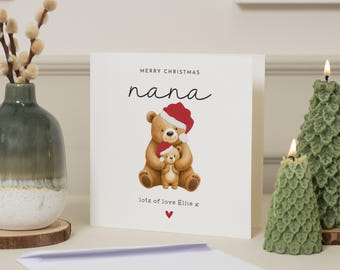 May include: A Christmas card with a teddy bear illustration. The card reads "Merry Christmas Nana" with a heart and the words "lots of love Ellie x". Two green tree-shaped candles and a vase with decorative branches are in the background.