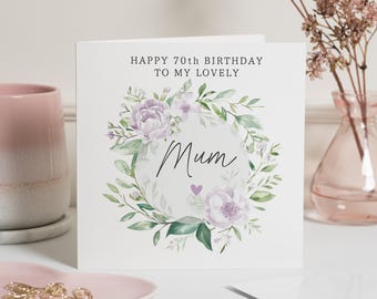 May include: A white greeting card with the text "HAPPY 70TH BIRTHDAY TO MY LOVELY Mum" in black. The card features a floral wreath with lavender flowers and green leaves. A pink vase and a pink mug are in the background.