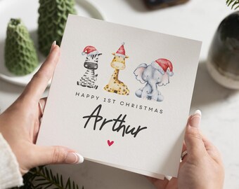 Op de afbeelding: Vierkante wenskaart met aquarel illustraties van een zebra, giraf en olifant, elk met een kerstmuts. De kaart leest "HAPPY 1ST CHRISTMAS" en "Arthur" met een klein rood hart.