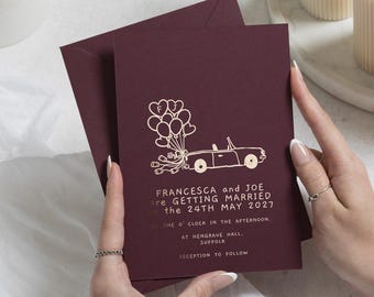 Invitaciones de boda con detalles dorados, invitaciones de boda color burdeos, invitación con dibujo a mano de un coche y un globo, invitación de boda divertida y personalizada.