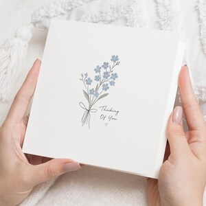 Può includere: Un biglietto d'auguri quadrato bianco con una delicata illustrazione di un piccolo mazzo di fiori non-ti-scordar-di-me azzurri legati con un sottile fiocco a tratto. Il testo Thinking Of You è stampato in un semplice carattere corsivo nero.