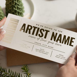 Könnte beinhalten: Ein cremefarbenes Konzertticket mit goldfarbenen Buchstaben und einem Barcode. Auf dem Ticket steht "WE'RE GOING TO SEE ARTIST NAME" und es gibt Platz für den Veranstaltungsort, das Datum und eine persönliche Nachricht. Gehalten von Händen.