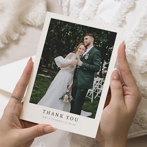 Peut inclure: Une personne tient une carte de remerciement crème. La carte présente une photo d'un couple de mariage. Le texte "THANK YOU" est imprimé en bas, avec les noms "AMY & THOMAS" et la date "21.06.26".