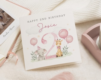 May include: A square birthday card with the text "Happy 2nd Birthday Josie" in black and pink. The card features illustrations of an elephant, giraffe, balloons, and the number 2. The card is on a white surface.