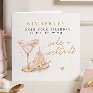 May include: A cream-coloured birthday card with gold text reading "Kimberley" and "I hope your birthday is filled with cake + cocktails." The card features a watercolour illustration of a cocktail and cake.
