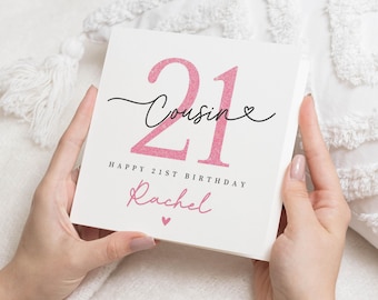 May include: A white birthday card held by hands, featuring the number "21" in pink glitter, with the word "Cousin" in cursive. The card also includes the text "Happy 21st Birthday Rachel" and a small pink heart.