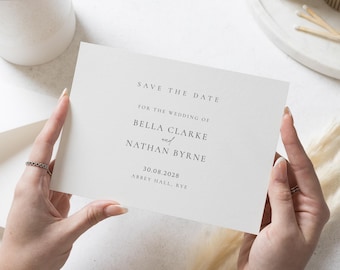 Könnte beinhalten: Eine cremefarbene Hochzeitseinladungskarte mit dem Text "SAVE THE DATE" und Hochzeitsdetails für Bella Clarke und Nathan Byrne am 30.08.2028 in Abbey Hall, Rye. Die Karte wird von Händen gehalten und hat ein minimalistisches Design.