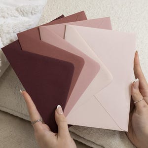 Może przedstawiać: Zestaw pięciu papierowych kopert ułożonych w wachlarz, prezentujących gradient kolorów od głębokiego burgundu i ciemnego kasztanu po przybrudzony róż, jasny wrzos i blady pudrowy róż, wszystkie z gładkim, matowym wykończeniem i spiczastymi klapkami.