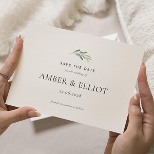 Op de afbeelding: Een crèmekleurige huwelijksuitnodiging met de tekst "SAVE THE DATE" en de namen "AMBER & ELLIOT" in het zwart gedrukt. De datum "22.06.2028" wordt ook weergegeven. De uitnodiging heeft een groen bladerontwerp en de woorden "formal invitation to follow".