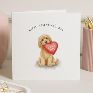 May include: A Valentine's Day card featuring a light brown dog holding a red heart with the words "Love You." The card has the text "Happy Valentine's Day" at the top. The card is on a white surface.