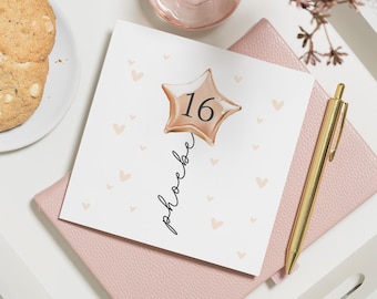 May include: A white card with a gold star balloon with the number 16 on it. The card has the words "Happy 16th" written on it in black cursive. The card is surrounded by small pink hearts.