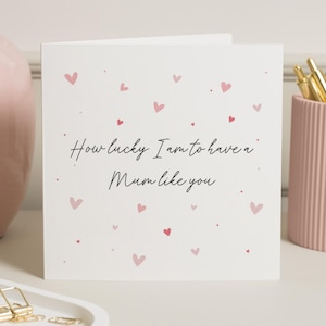 Könnte beinhalten: Eine weiße Grußkarte mit dem Text "How lucky I am to have a Mum like you" in schwarzer Schrift. Verziert mit kleinen rosa Herzen. Im Hintergrund sind ein rosa Stifthalter und eine rosa Vase zu sehen.