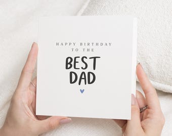 May include: A white greeting card with the text "HAPPY BIRTHDAY TO THE BEST DAD" in black font. A small blue heart is below the text. The card is held by a person.