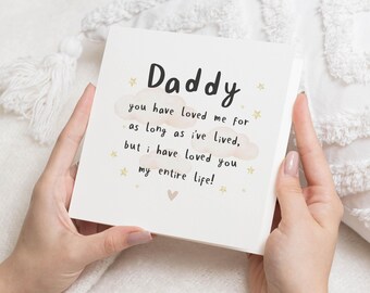 May include: A square, off-white card with the word "Daddy" in black script. The card features a message in black text with cloud and star illustrations. The card is held by hands against a soft, white background.