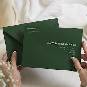 Op de afbeelding: Een donkergroene envelop en bijpassende uitnodigingskaart met witte bedrukte tekst. De kaart toont de namen LUCY & MAX CARTER, een adres in Newcastle, Engeland, en een sectie met het label KINDLY REPLY TO met adresgegevens.