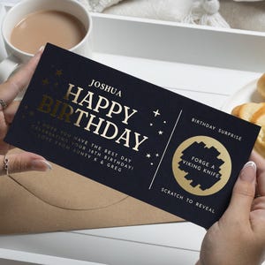 May include: A navy blue birthday scratch card with gold text reading "Joshua Happy Birthday". The card features the text "Forge a Viking Knife" and "Scratch to Reveal". The card is held over a brown envelope.