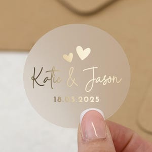 Könnte beinhalten: Ein runder, mattierter Aufkleber mit goldfarbenem Text und zwei Herzen. Der Text lautet "Katie & Jason" über dem Datum "18.05.2025". Der Aufkleber wird von einer Person mit hellrosa Maniküre gehalten.