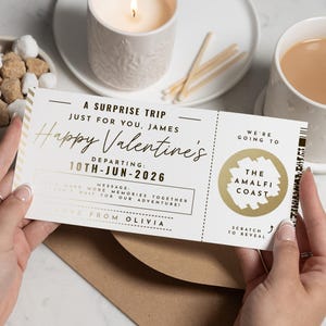 May include: A Valentine's Day surprise trip ticket with gold foil detailing. The ticket says "Happy Valentine's" and "The Amalfi Coast." The departure date is 10th June 2026. The ticket is held by a person.