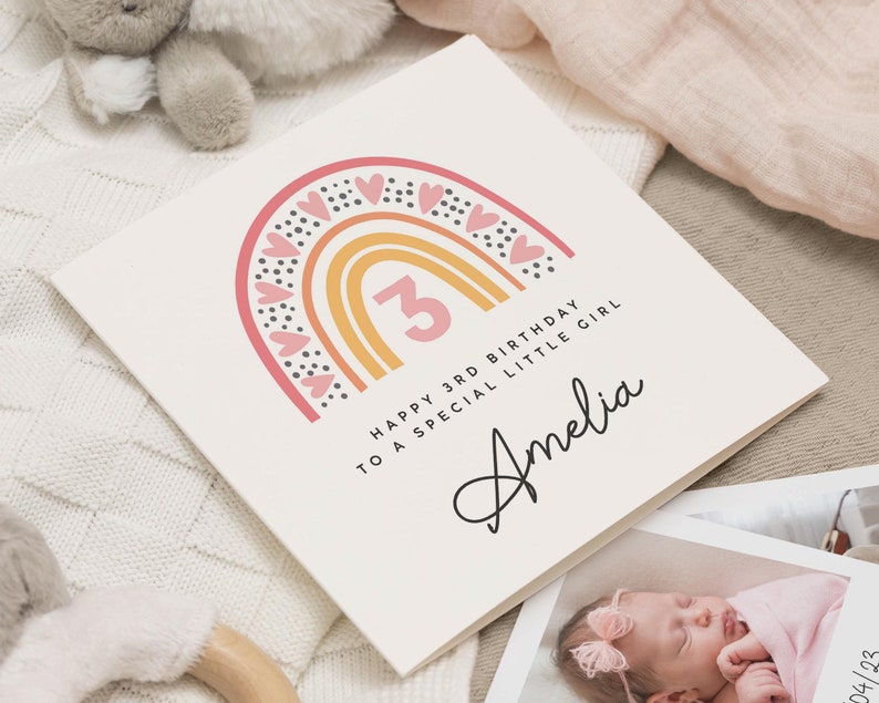 Op de afbeelding: Een roze en gele regenboog met zwarte stippen en roze harten. Het getal 3 staat in het midden van de regenboog. De tekst "Happy 3rd Birthday to a special little girl Amelia" staat onder de regenboog.
