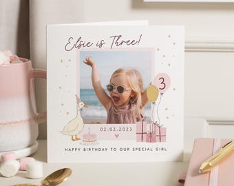 May include: A square birthday greeting card featuring a central photo of a child with arms raised, surrounded by illustrations of a duck in a party hat, a birthday cake, balloons with the number three, and gifts. Text reads Elsie is Three!