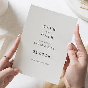 Op de afbeelding: Een crèmekleurige 'Save the Date'-kaart, vastgehouden door handen, met elegante zwarte tekst. De kaart leest 'SAVE the DATE' boven 'for the wedding of LAURA & KYLE' en de datum '22.07.28'. De zin 'formal invitation to follow' staat onderaan.