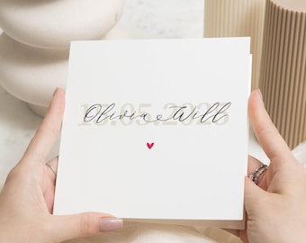 Op de afbeelding: Een witte kaart met de tekst "Olivia & Will" in zwarte cursieve letters en een klein rood hartje.