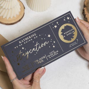 May include: A navy blue gift card with gold foil lettering and star details. The card says "Katherine, we're going on a staycation" with a scratch-off area for a weekend in Dublin. Includes "Love from Mum & Nanna" and dates.