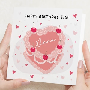 Könnte beinhalten: Eine Geburtstagskarte, die von Händen gehalten wird, mit einem herzförmigen Kuchendesign, Kirschen und dem Namen "Anna". Die Karte lautet "Happy Birthday Sis!" mit verstreuten rosa Herzen.