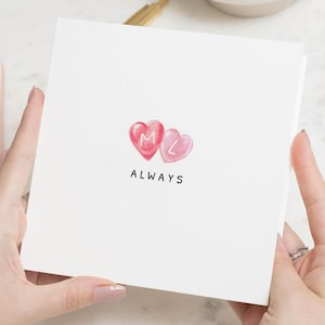 May include: A white greeting card featuring two watercolor hearts, one pink and one red, with the letters "M" and "L" inside. The word "ALWAYS" is written in black below the hearts. The card is held by a person.