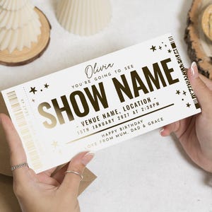 Puede incluir: Una persona sostiene un boleto blanco con texto dorado y detalles de estrellas. El boleto dice "SHOW NAME" en letras grandes, con detalles que incluyen el lugar, la fecha y la hora. El boleto también incluye el nombre "Olivia" y un mensaje de cumpleaños.