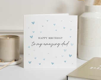 May include: A square white greeting card featuring small light blue hearts scattered across the front. The text reads HAPPY BIRTHDAY in a bold sans-serif font, followed by the script text to my amazing dad in black ink.