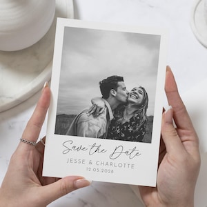 May include: A black and white photograph of a couple kissing, printed on a white card. The text "Save the Date" is written in cursive, with the names "Jesse & Charlotte" and the date "12.05.2028" below. The card is held by a person.