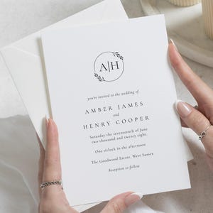 May include: A white wedding invitation with black text, held by hands. The invitation features a circular design with the initials "A" and "H" and the words "you're invited to the wedding of Amber James and Henry Cooper".