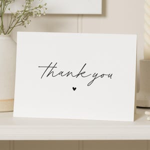 Può includere: Un biglietto d'auguri rettangolare bianco con il testo thank you in un carattere corsivo nero centrato sulla parte anteriore. Sotto il testo si trova una piccola icona a forma di cuore nero. Il biglietto è realizzato in carta liscia.