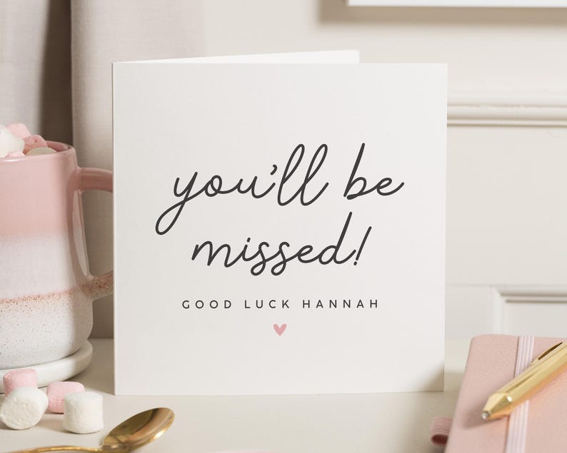 May include: White card with black text that reads "you'll be missed!" and "GOOD LUCK HANNAH" with a small pink heart below the text.