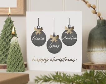 May include: A white card with a gold inscription that reads "happy christmas". Three black ornaments with gold bows hang from the top of the card. Each ornament has a name written on it: Olivia, Lewis, and Amelia.