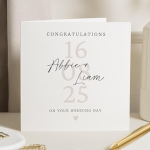 Op de afbeelding: Een vierkante huwelijkskaart met de tekst "Gefeliciteerd" bovenaan. De namen "Abbie + Liam" zijn in cursief geschreven, met de cijfers "16 08 25" eronder. De woorden "On Your Wedding Day" staan onderaan, met een klein hartje.