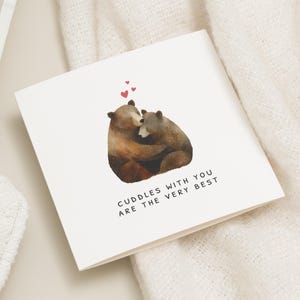 May include: A white greeting card featuring a watercolour illustration of two brown bears embracing. Above the bears are three small red hearts. The text reads "Cuddles with you are the very best".