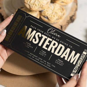 May include: A black and gold Amsterdam boarding pass with the text "PACK YOUR BAGS! WE'RE GOING TO... AMSTERDAM". The pass includes the date, time, and gate information. The pass is held in front of a brown envelope.
