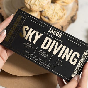 Könnte beinhalten: Ein schwarzes und goldenes Fallschirmspringer-Ticket mit dem Text "SKY DIVING" in großen Buchstaben. Das Ticket enthält den Namen des Empfängers, Datum, Uhrzeit und Ortsangaben. Das Ticket wird vor einem braunen Umschlag gehalten.