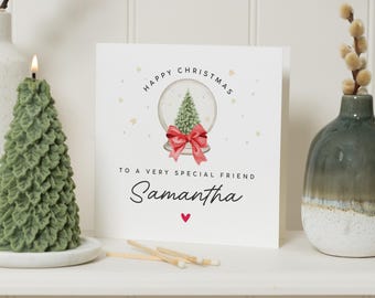 May include: A Christmas card with a snow globe design, a red bow, and the text "Happy Christmas to a very special friend Samantha". A green tree-shaped candle and a vase with decorative stems are on the sides. Matches are in the foreground.