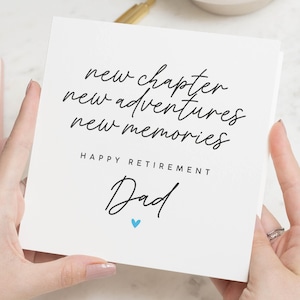 May include: A white square card with black script that reads "new chapter, new adventures, new memories." Below, it says "HAPPY RETIREMENT Dad" with a small blue heart. The card is held by two hands.